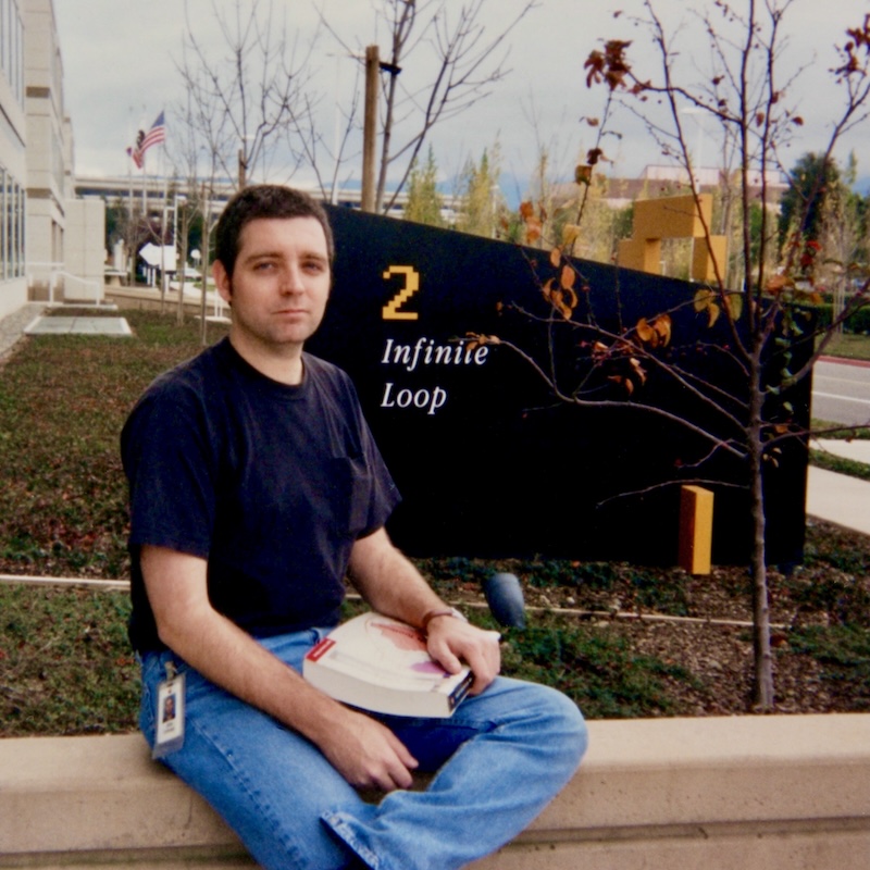 Man sitting outside 2 Infinite Loop, Apple’s former headquarters in Cupertino, holding a book with an ID badge clipped to his jeans.